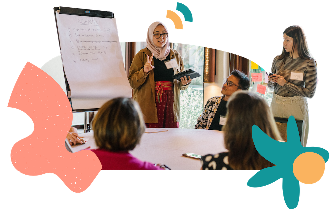 A facilitator standing in front of a flipchart, facing and speaking to a group sitting around a table