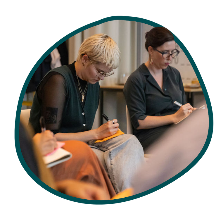 Two women sit next to each other writing in their notebooks