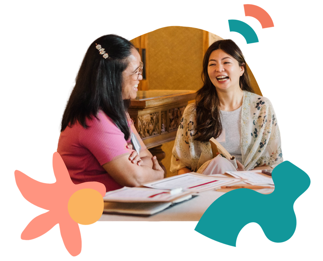 Two women are sitting at a table facing each other and laughing