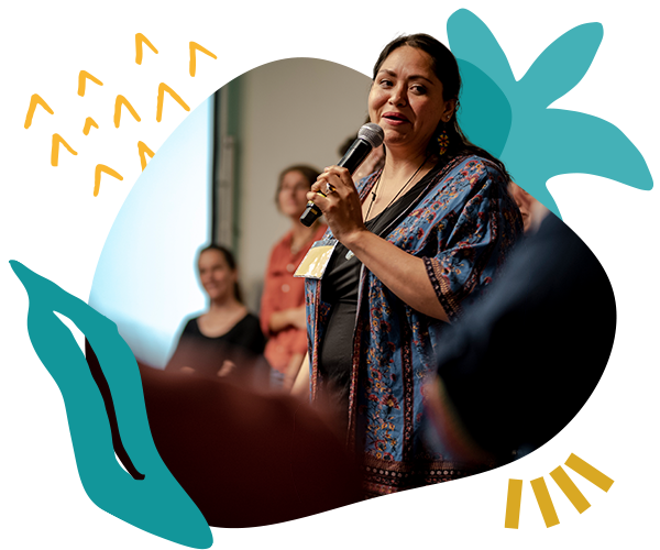 A woman stands speaking into a microphone in front of a room of people during a training