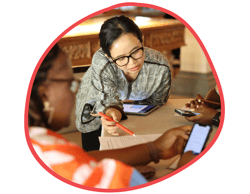 A woman with glasses is sitting at a table with others, looking at a piece of paper with a pen as the others research on their cellphones