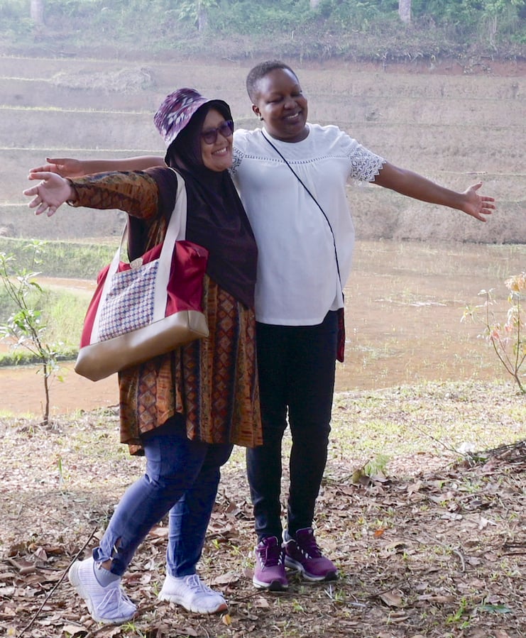 Two women are standing together with their arms outstretched in front of a body of water in a forest area