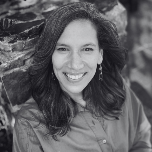 A portrait of Ella Scheepers posed outside by a large rock. She is wearing a spring green button-up shirt. She is smiling and wears her dark hair loose. She had dangling earrings.