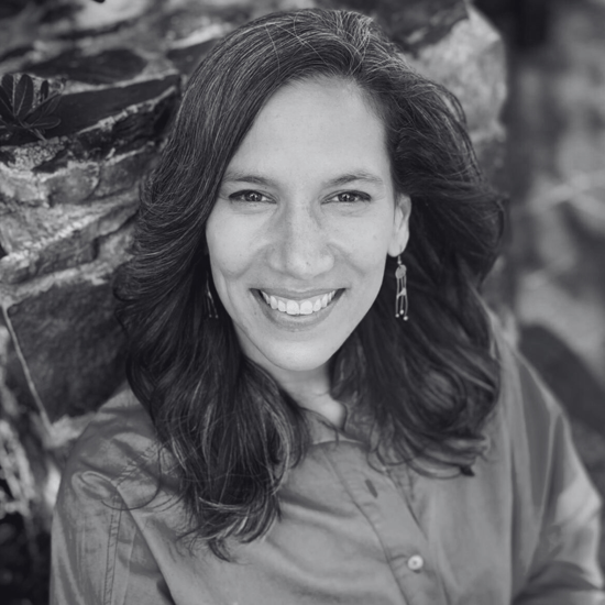 A portrait of Ella Scheepers posed outside by a large rock. She is wearing a spring green button-up shirt. She is smiling and wears her dark hair loose. She had dangling earrings.
