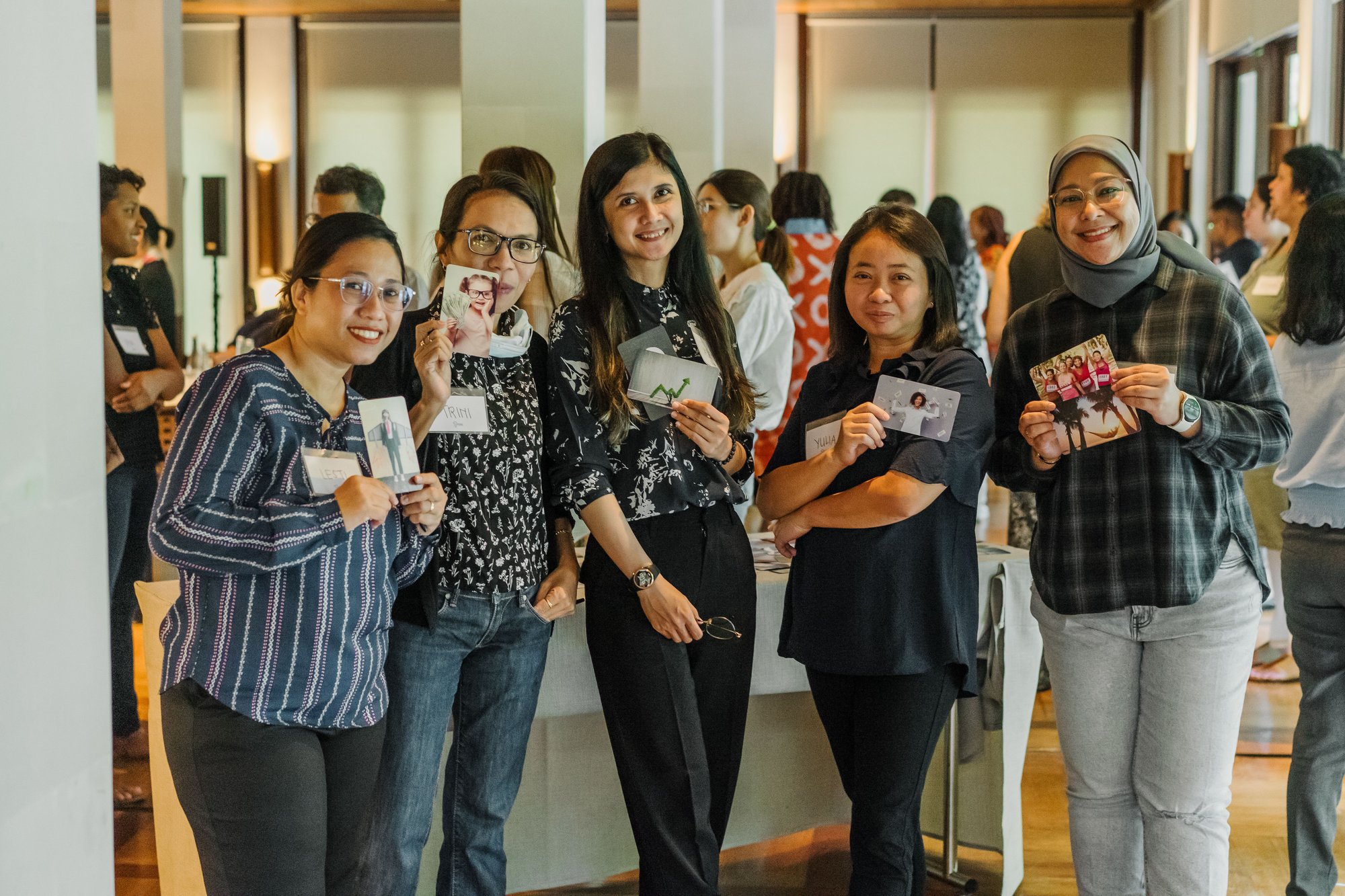 A group of women stand with cards representing their relationships with money. One smiles and wears a headscarf. The other four smile and hold up their cards.