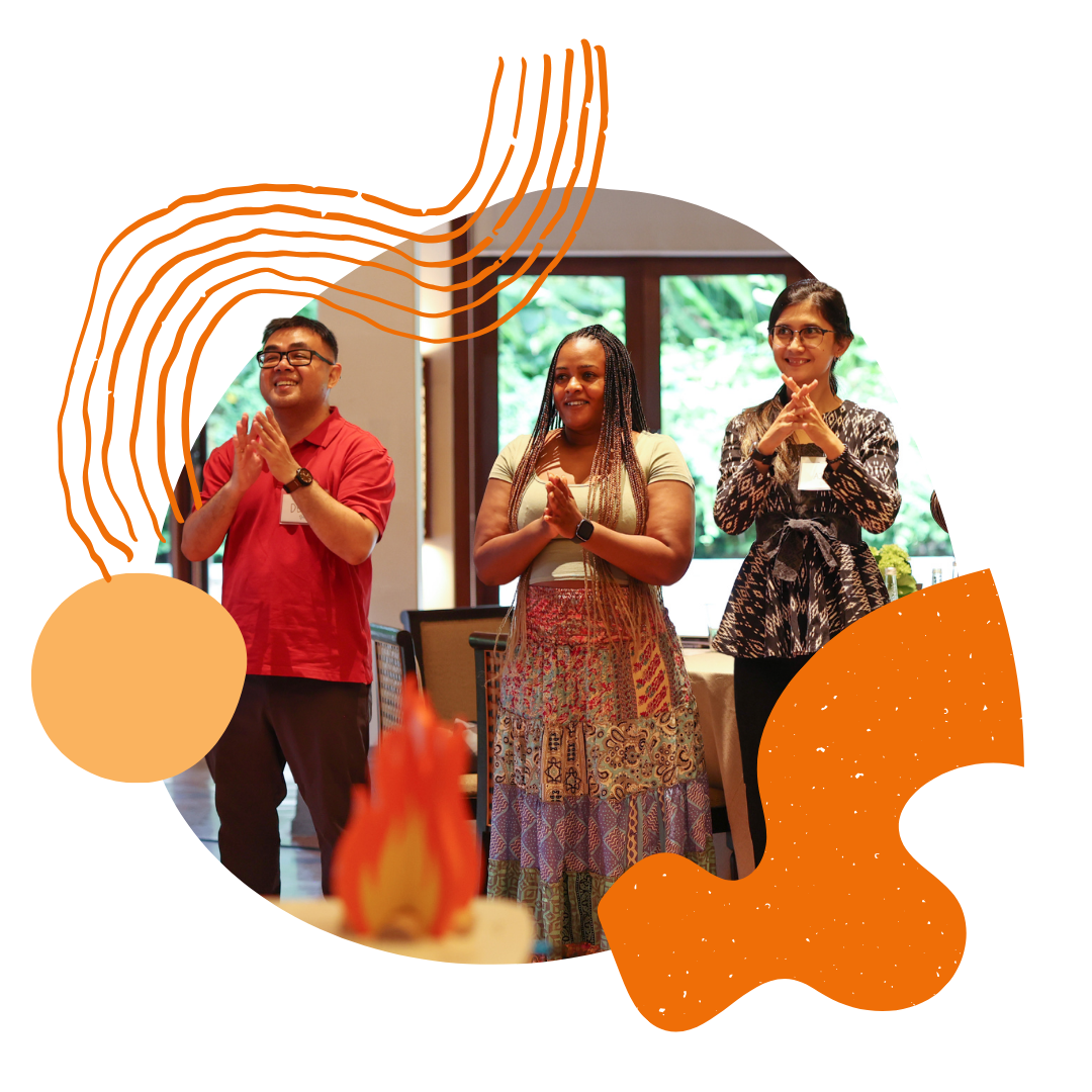 Three people stand together as part of a circle, smiling and with their hands clasped, behind a wooden fire