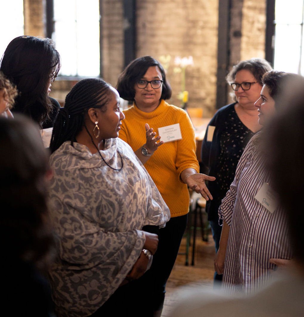 A group of women stand in a circle having a discussion during a training and participants are all paying attention to a woman in the center with a yellow sweater