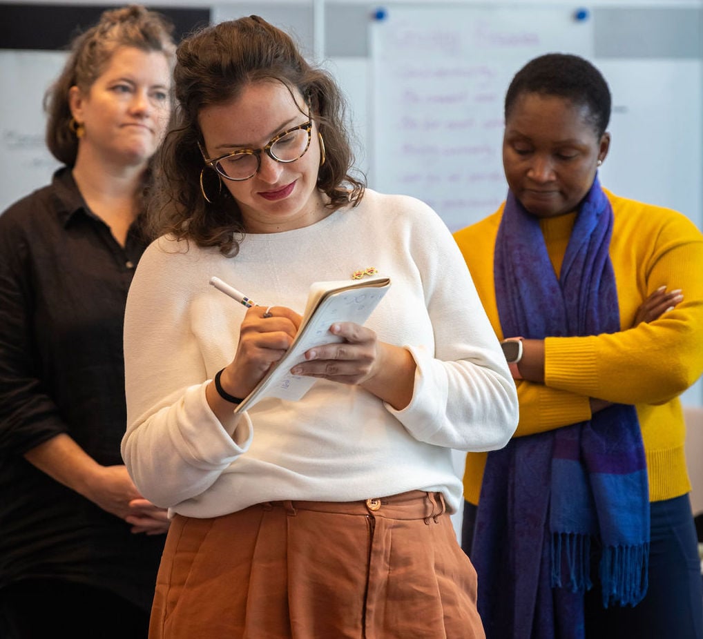 Three women standing with one at the forefront writing on a notepad