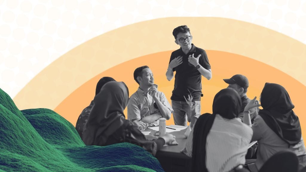 Group of people sitting around a table listening to a man standing in the middle