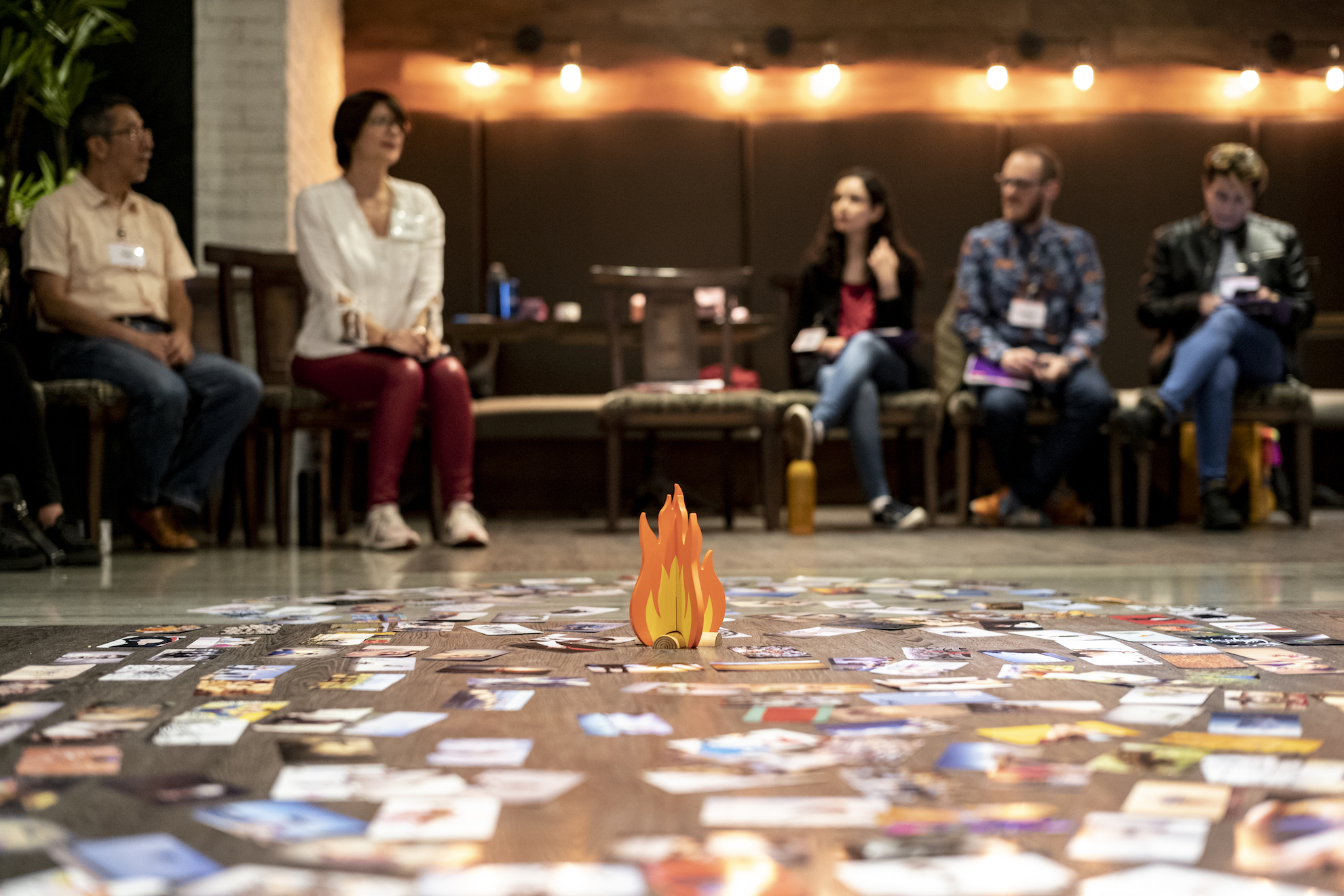 People are seated in a circle around dozens of cards scattered around the floor surrounding a fire