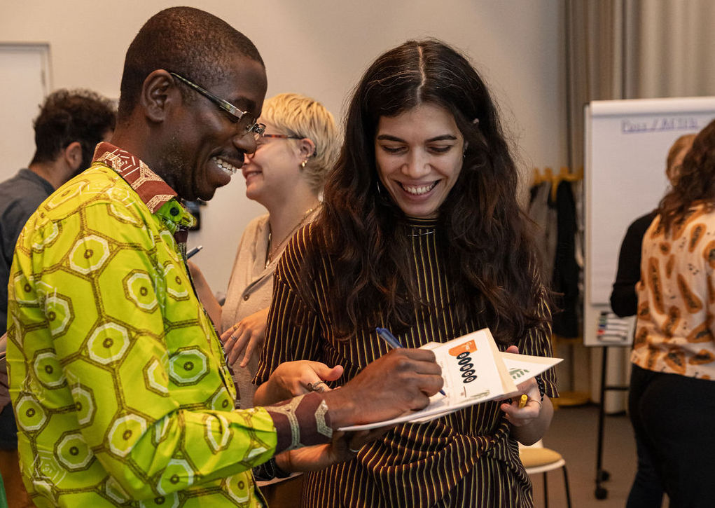 Two people stand next to each other during a training smiling and looking down as they write on a sheet of paper